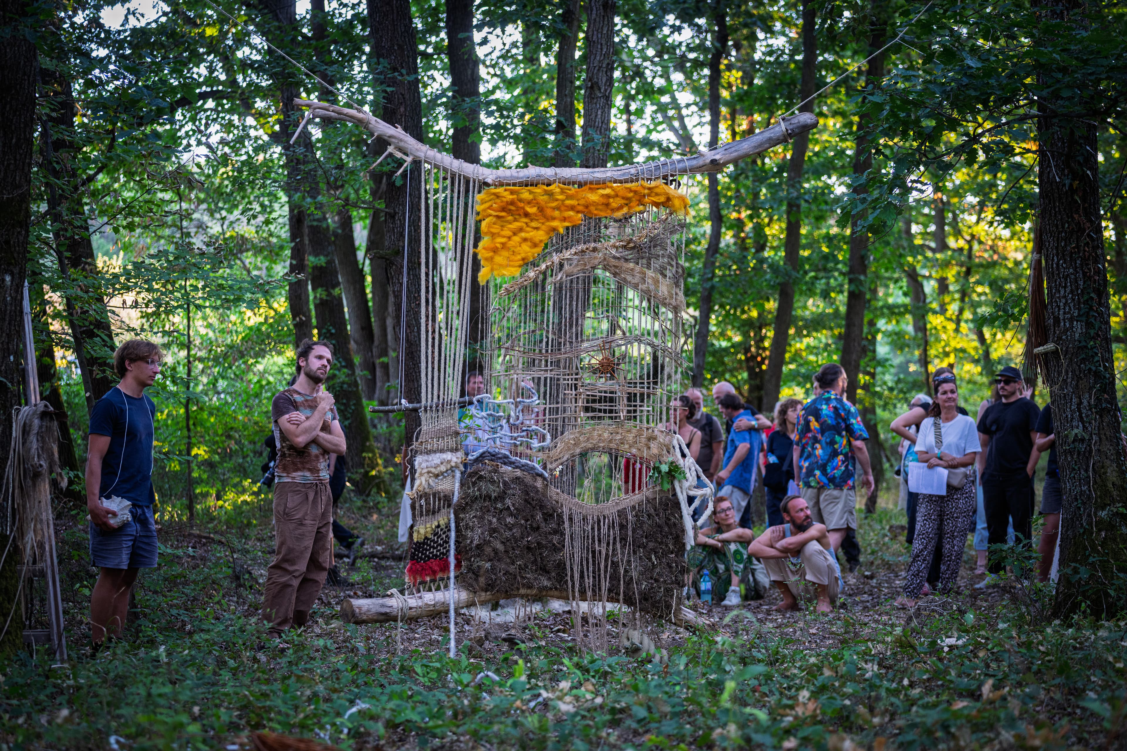 a handcrafted loom created from wood hung on two trees, woven with various textiles and organic materials. surrounded by a group of people in a forest. 
