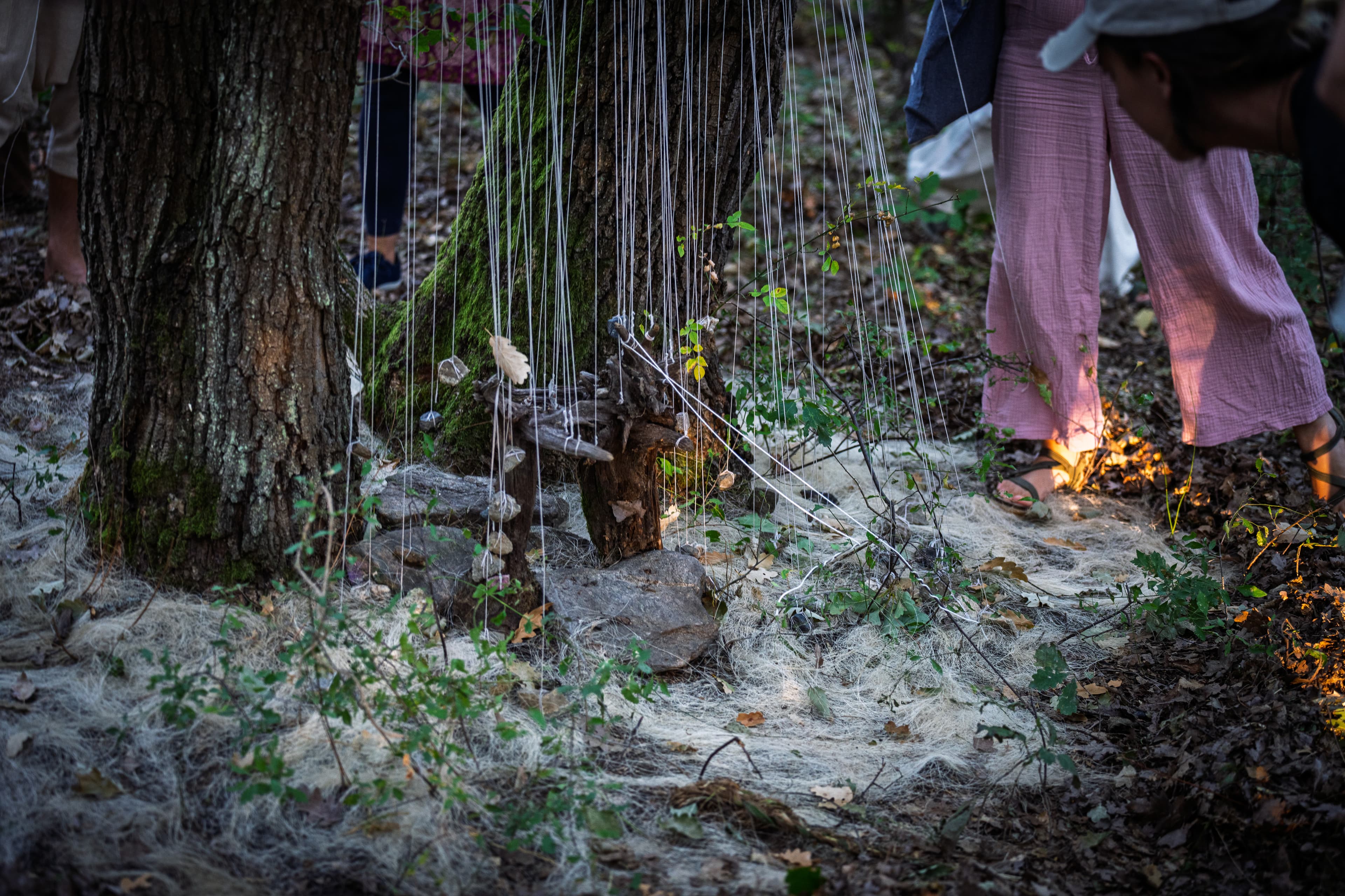 an altar made of wood, braided with threads where stones are hanging. situated next to a tree and forest floor.