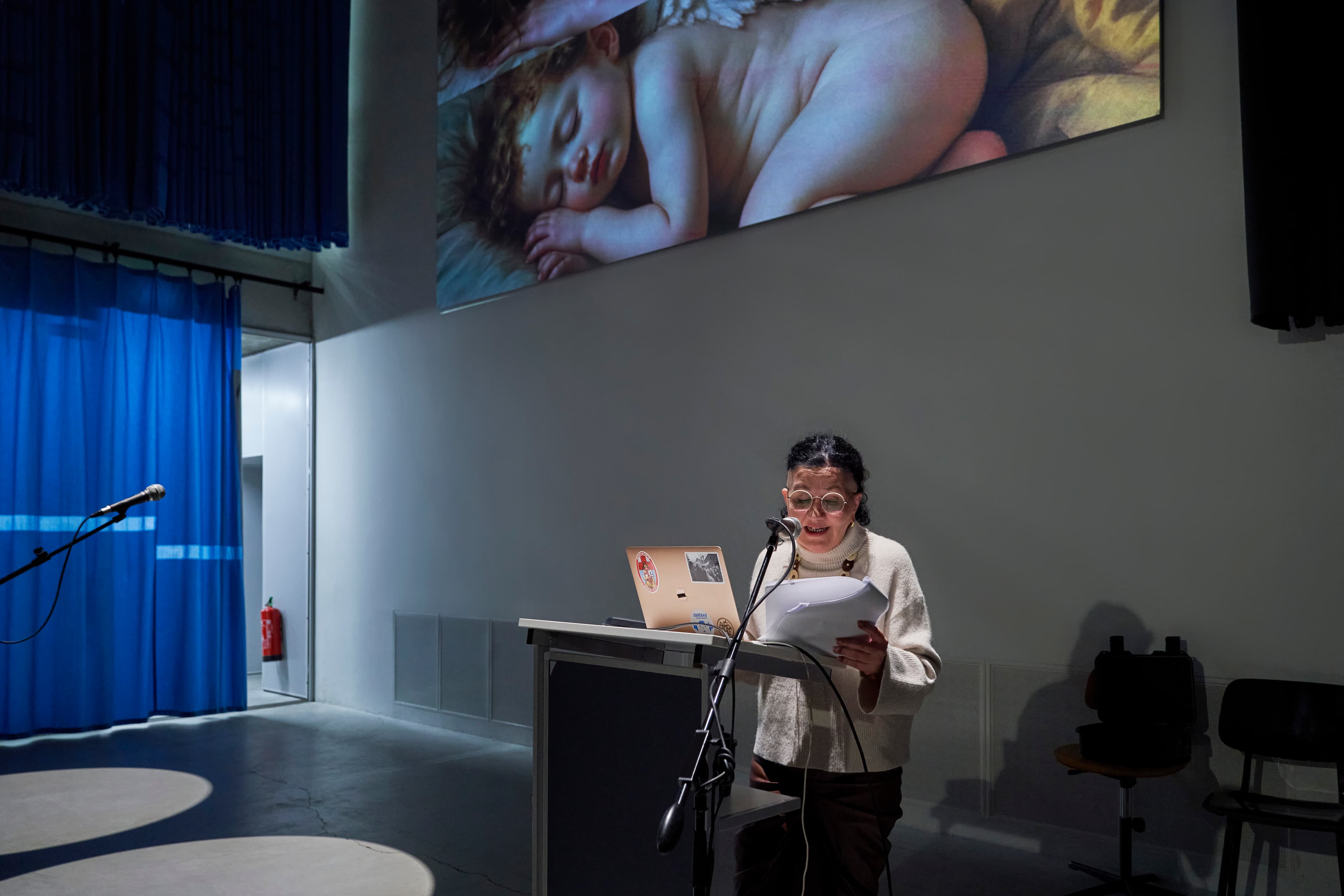 The image shows a women wearing white long jumper reading notes and giving a presentation in a darkly lit presentation space.