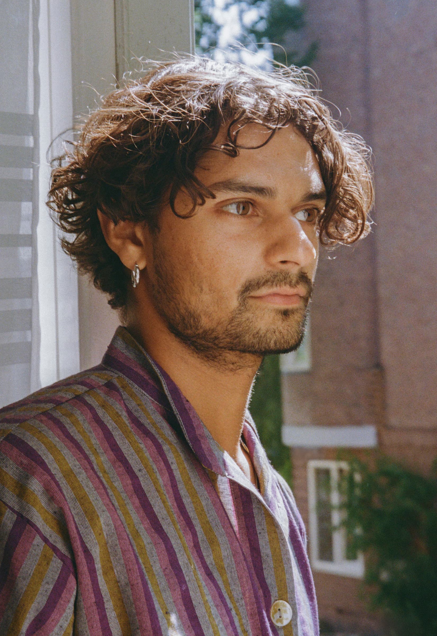 A portrait photograph of Kelvin, shown from the chest up, looking out of a window during summer. Kelvin wears a multicolored garment in shades of purple, lilac, mustard yellow, and grey. They have long, curly black hair that falls just below the ears and wear woven silver earrings.