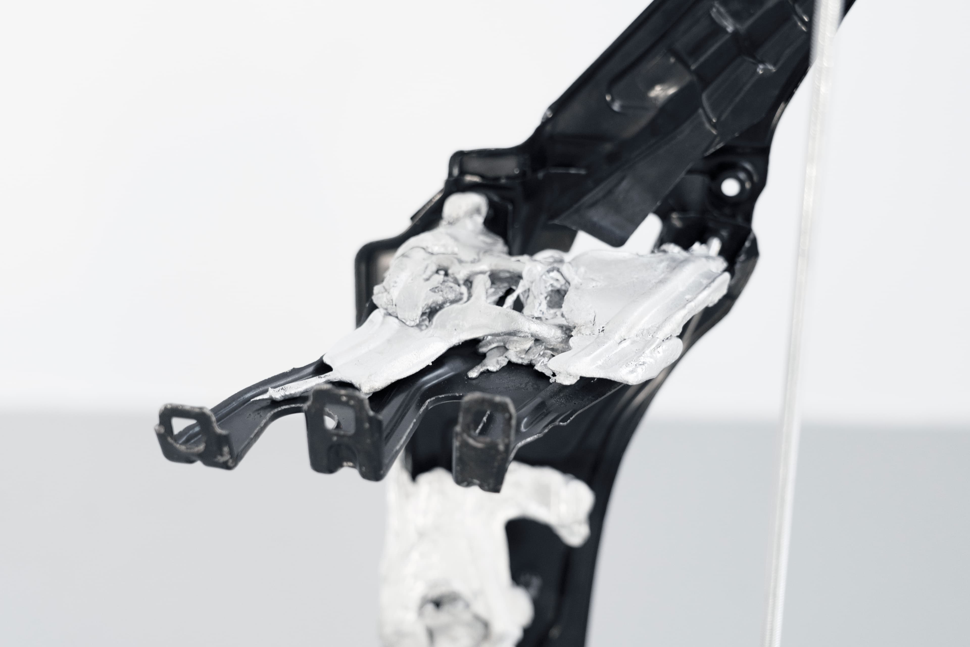 A close-up of a sculptural detail: solidified aluminium cast over a black automotive body panel, with an aluminium rod visible on the right.