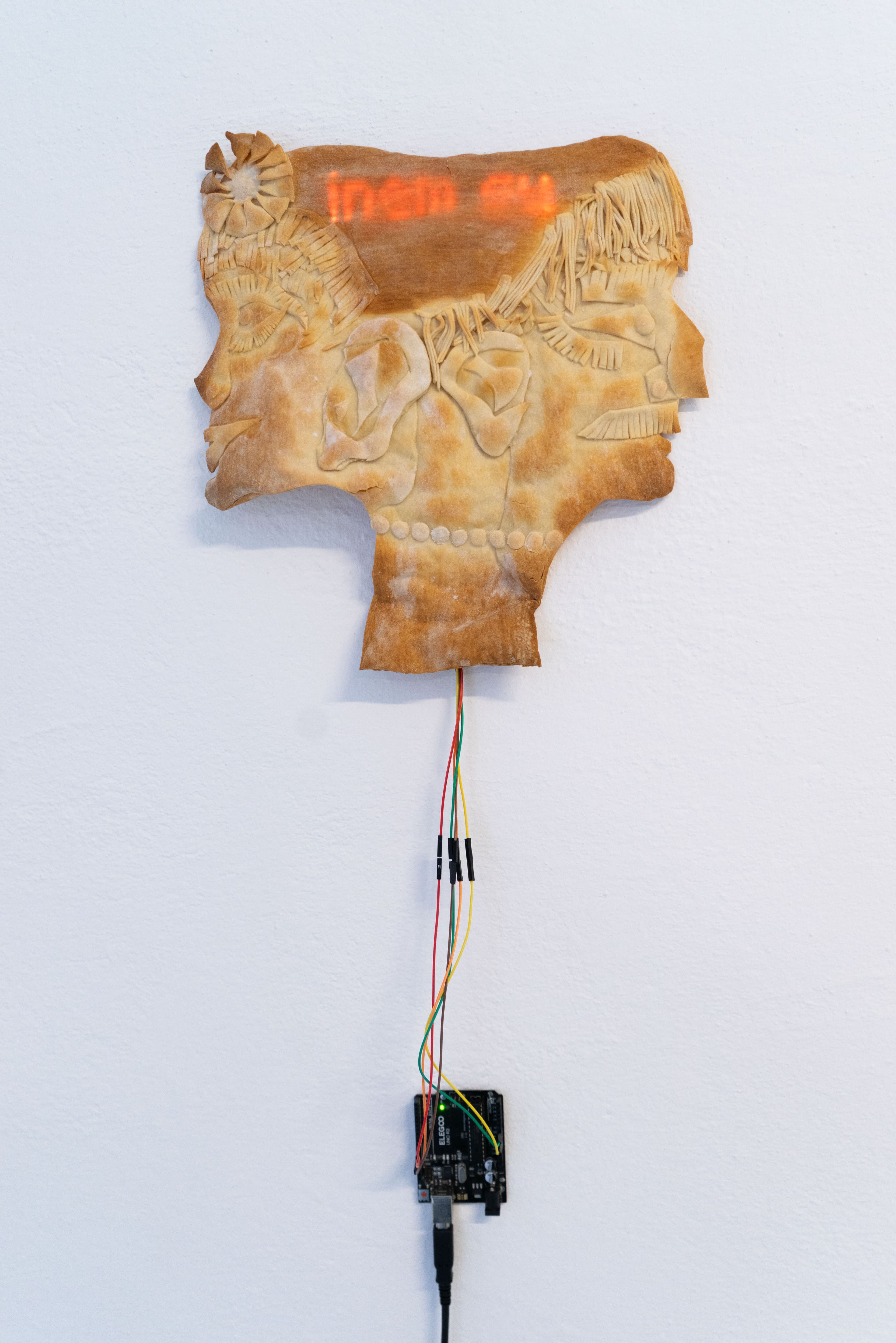 A double-faced mask made from bread, wires sticking out the bottom and a small LED panel that shines sentences through the thin layer of bread.