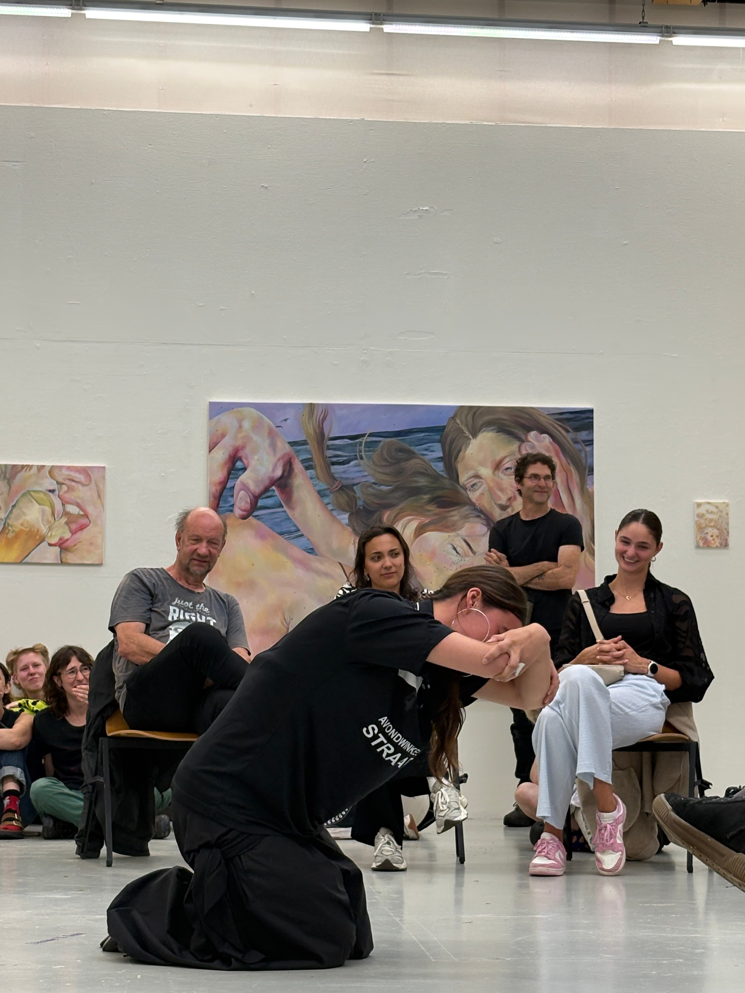 Female performer kneeling on the floor, wrapping her arms around each other, the public sits around her, laughing