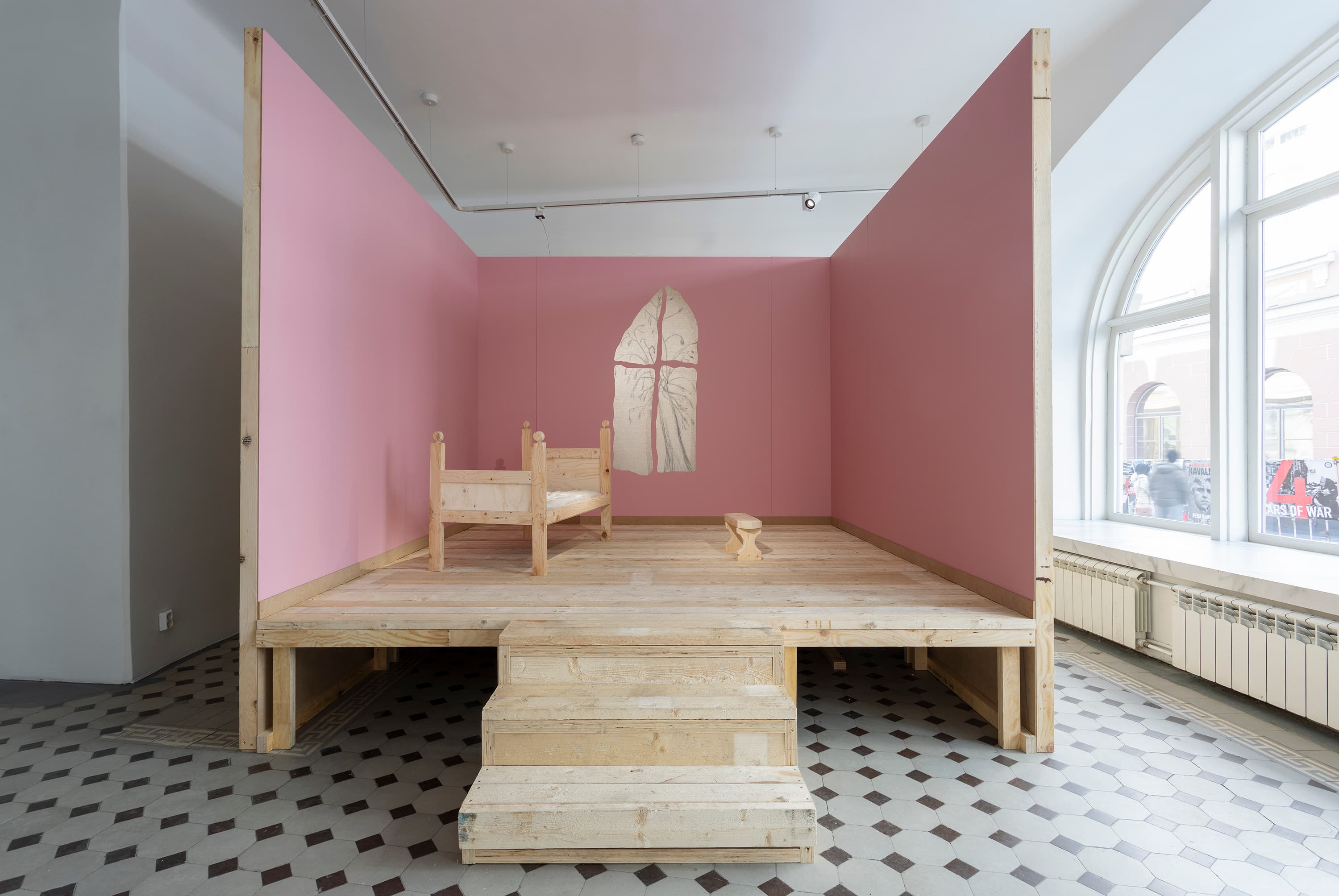 a square stage-like structure with three pink walls and a small wooden stair with three steps. one of the walls has a window-like hole, behind which is a drawing of a tree. on the stage, a very small bench and next to it, a small wooden bed.