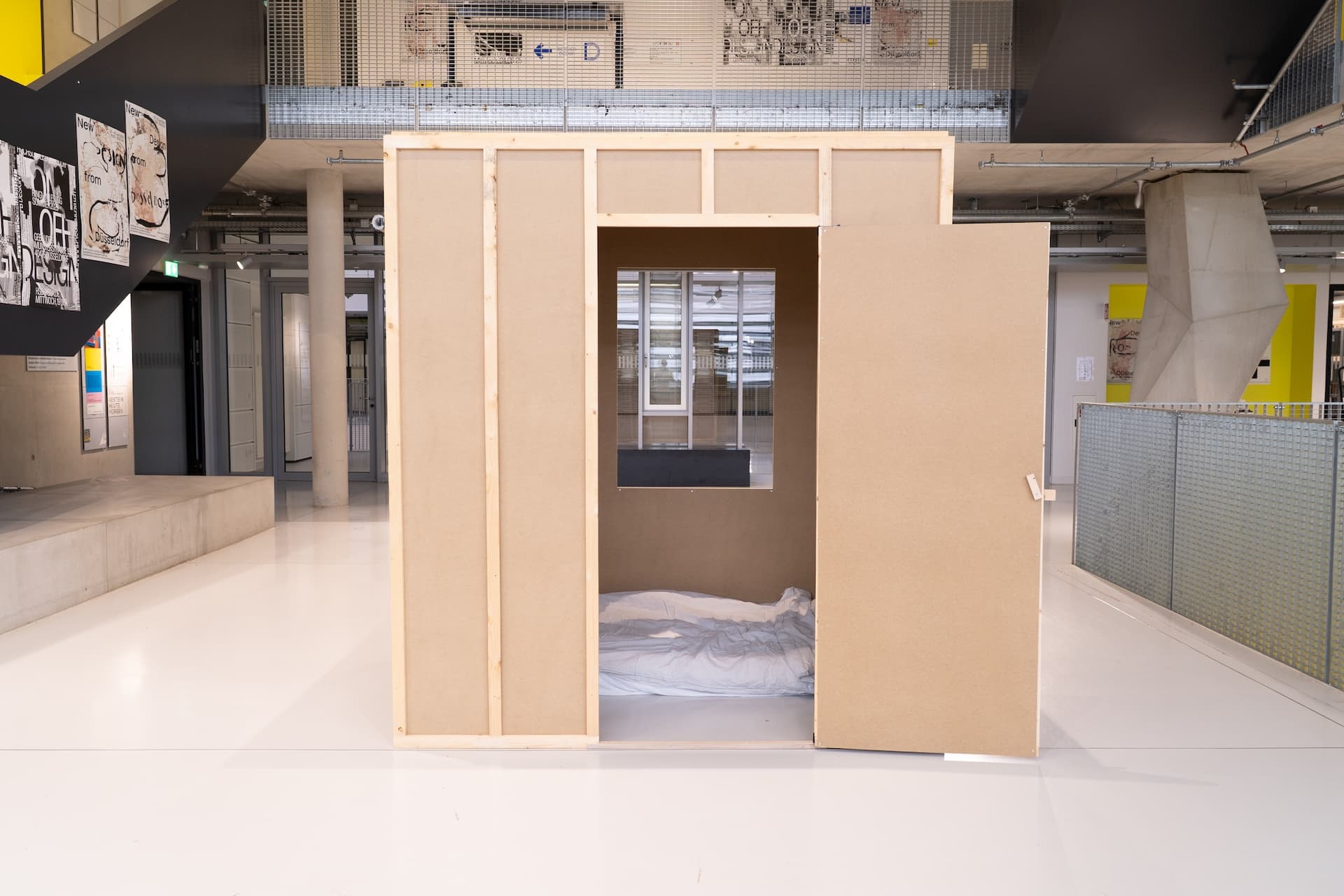 picture of a room box, 2 meters wide and 1,62 meters long in an open space in a public school. the rooms door is off-center to the right and open. inside the room a mattress is visible. 