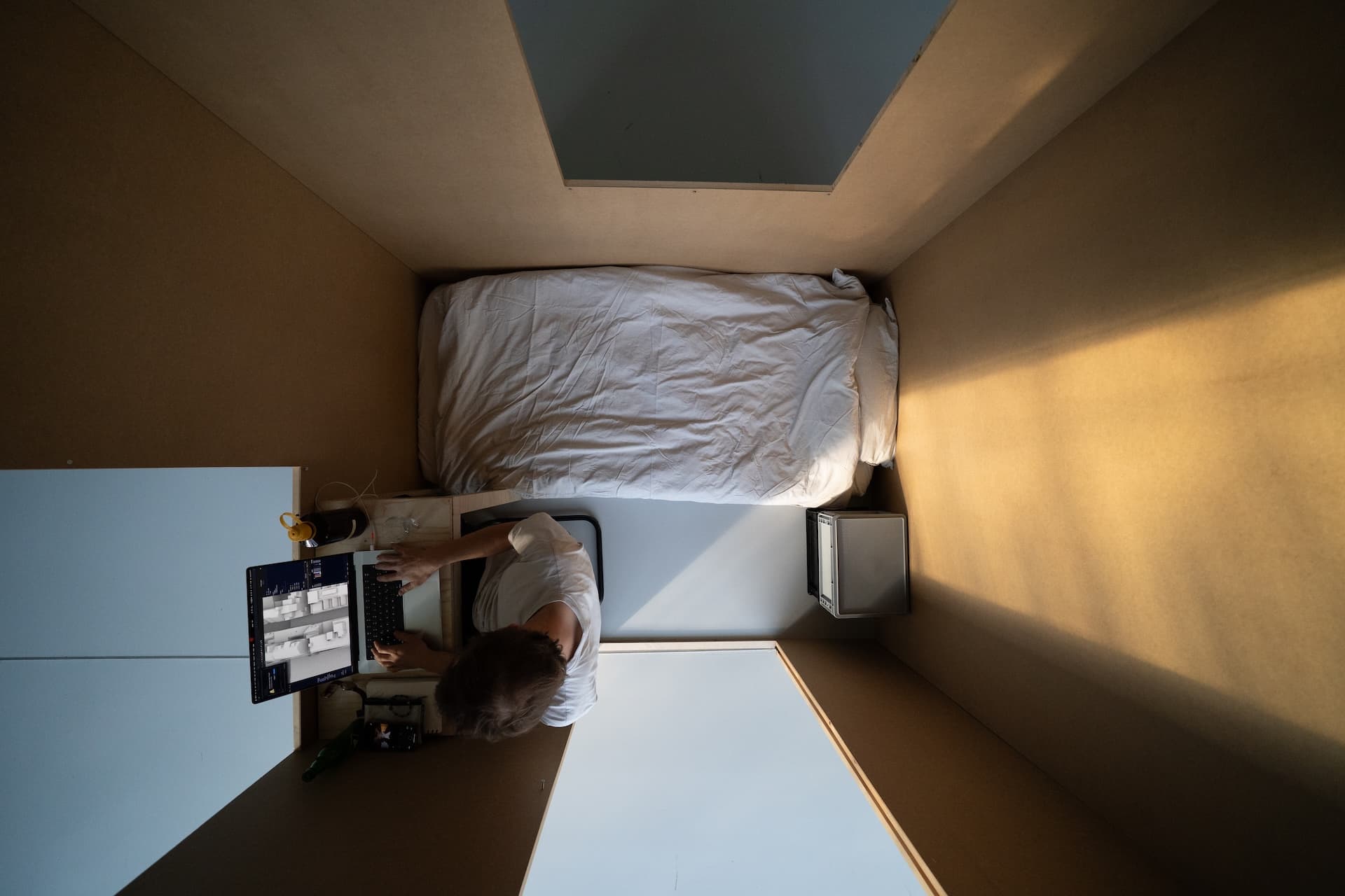 inside of a room that is 2 x 1,62 meters. objects in the room: a mattress, desk, chair, box. person sitting at the desk with a laptop. 