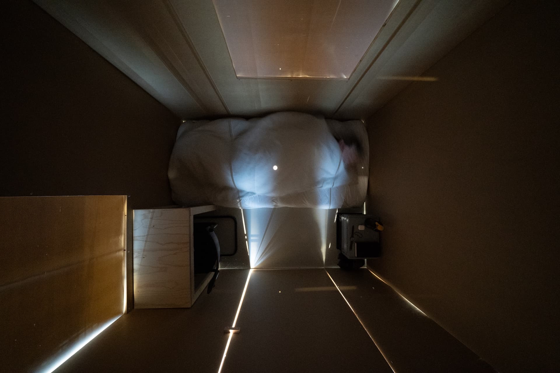 inside of a room that is 2 x 1,62 meters. the room is dark, some stray light here and there. objects in the room: a mattress, desk, chair, box. a person laying on the mattress