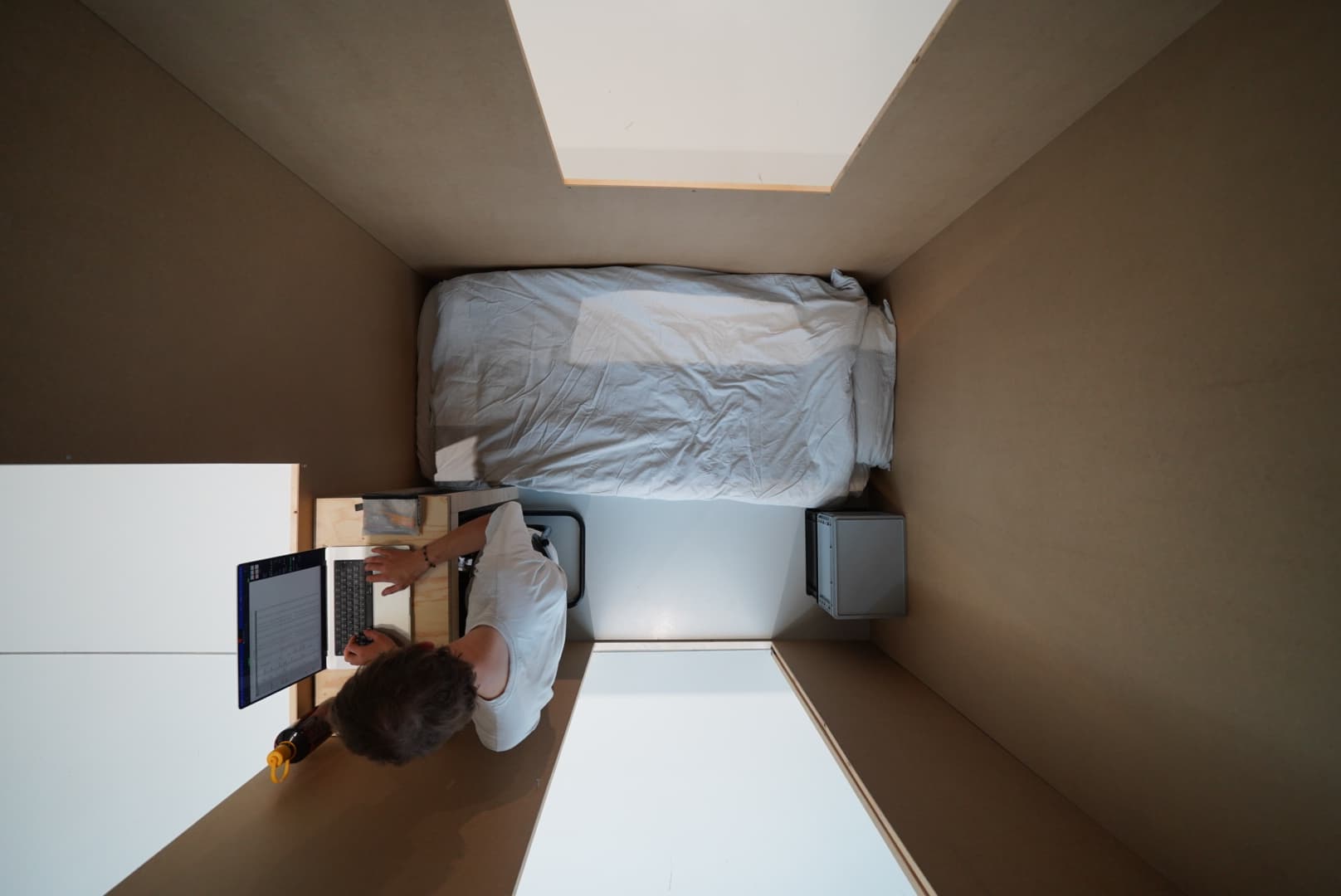 wide-angle picture of a small room taken from the ceiling. in the room are a bed, two boxes, a person on a desk. the room has three openings. 