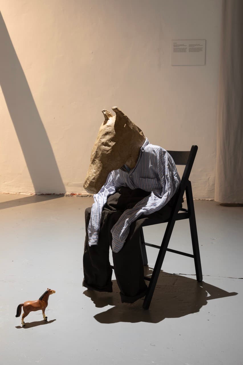 on the ground, a small plastic toy horse. sitting in a chair and looking down at the toy is a human body with a papier-mache horse head.