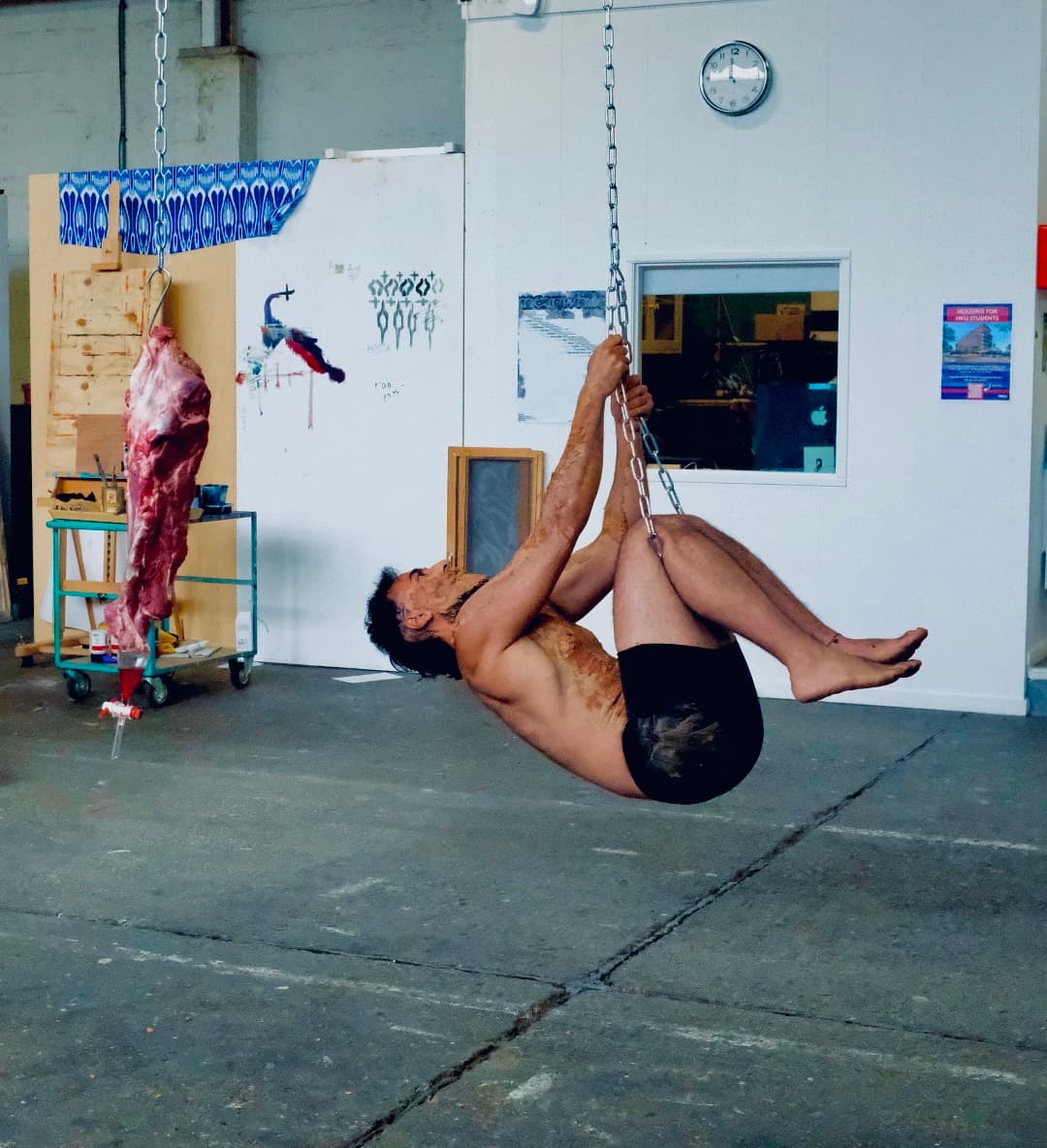 A visceral live performance confronting systemic displacement and bureaucratic control. Positioned beneath a suspended, bleeding cut of raw meat, the earth-coated artist mirrors the mechanics of slaughter by having their own legs hoisted by heavy chains. This "living sacrifice" transforms the tools of execution into a radical ritual of bodily sovereignty, asserting: No government owns my body