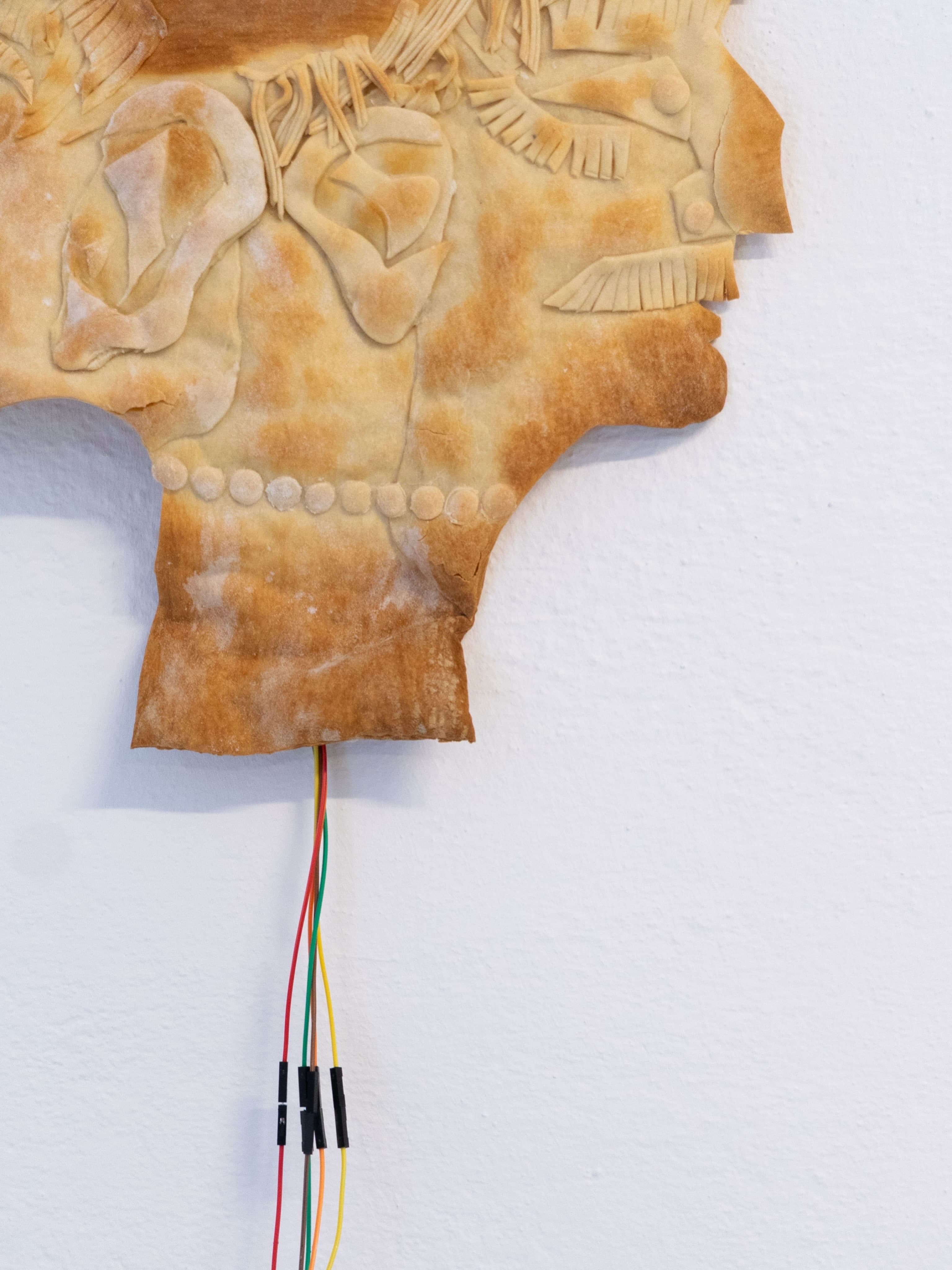 Closeup of a double-faced mask made from bread, wires sticking out the bottom and a small LED panel that shines sentences through the thin layer of bread.