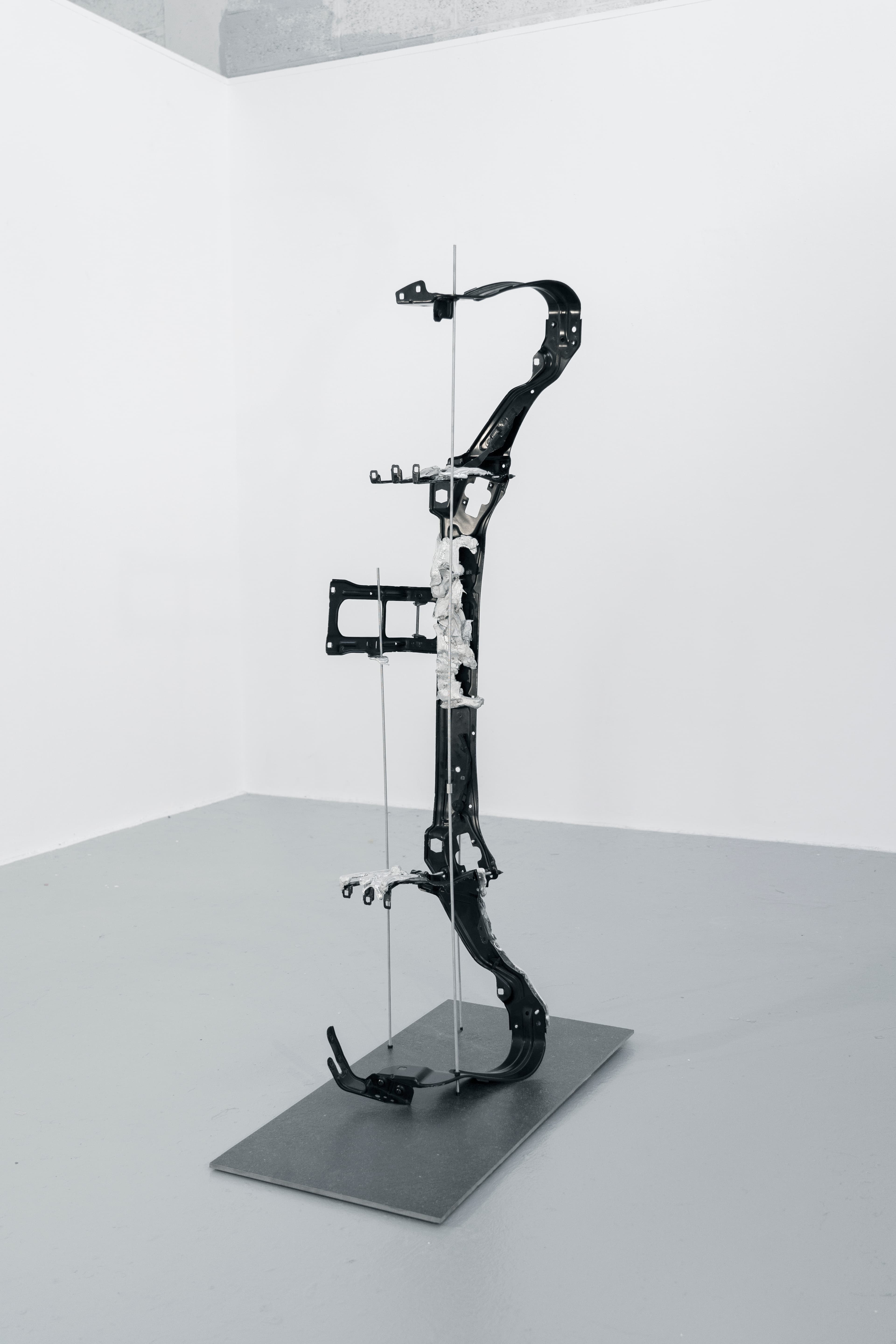 A tall vertical sculpture standing on a metal base. A black automotive body panel is threaded onto thin aluminium rods, with deformed white material at the centre.