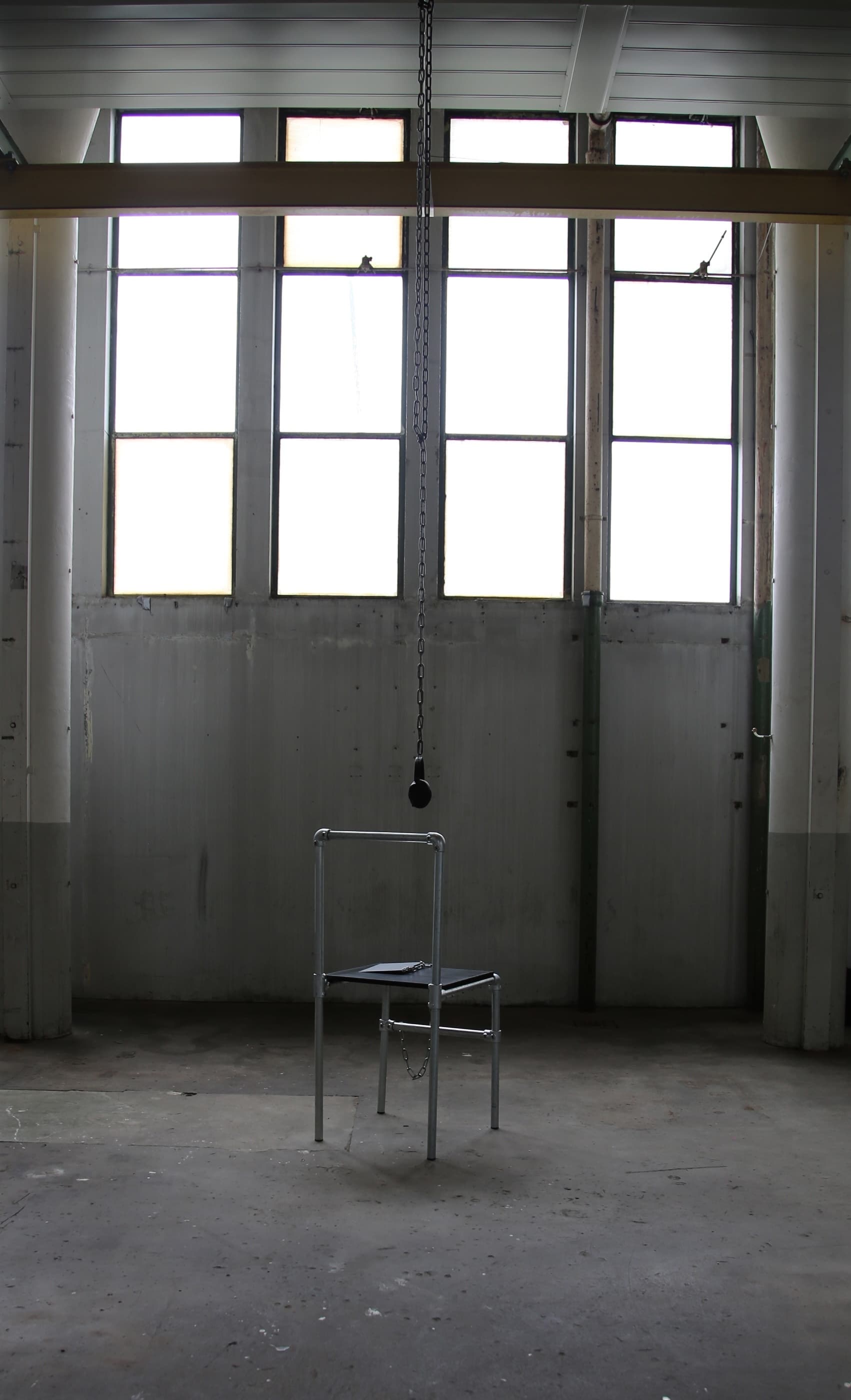 A steel chair facing away from the camera stands in the centre of a warehouse space. A pair of headphones hags down from the ceiling