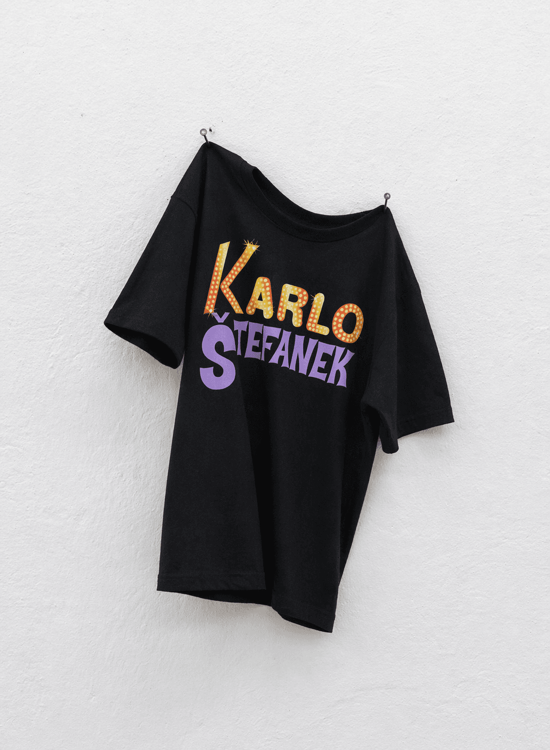 A black, short-sleeved T-shirt hangs loosely from two small metal nails pinned into a textured white gallery wall. The shirt is slightly tilted, with its fabric falling into natural folds and its hem hanging unevenly. Across the chest, a large, colourful graphic spells out “KARLO ŠTEFANEK” in two lines. “KARLO” appears in yellow, orange and red lettering decorated with small dots and sparkling star details, resembling illuminated marquee signs. Beneath it, “ŠTEFANEK” is printed in bold, playful purple lettering with exaggerated, irregular shapes. The vibrant, theatrical typography contrasts sharply with the black fabric and minimalist white background. Soft, even lighting highlights the garment’s texture and creates subtle shadows against the wall, giving the photograph the appearance of contemporary art documentation.