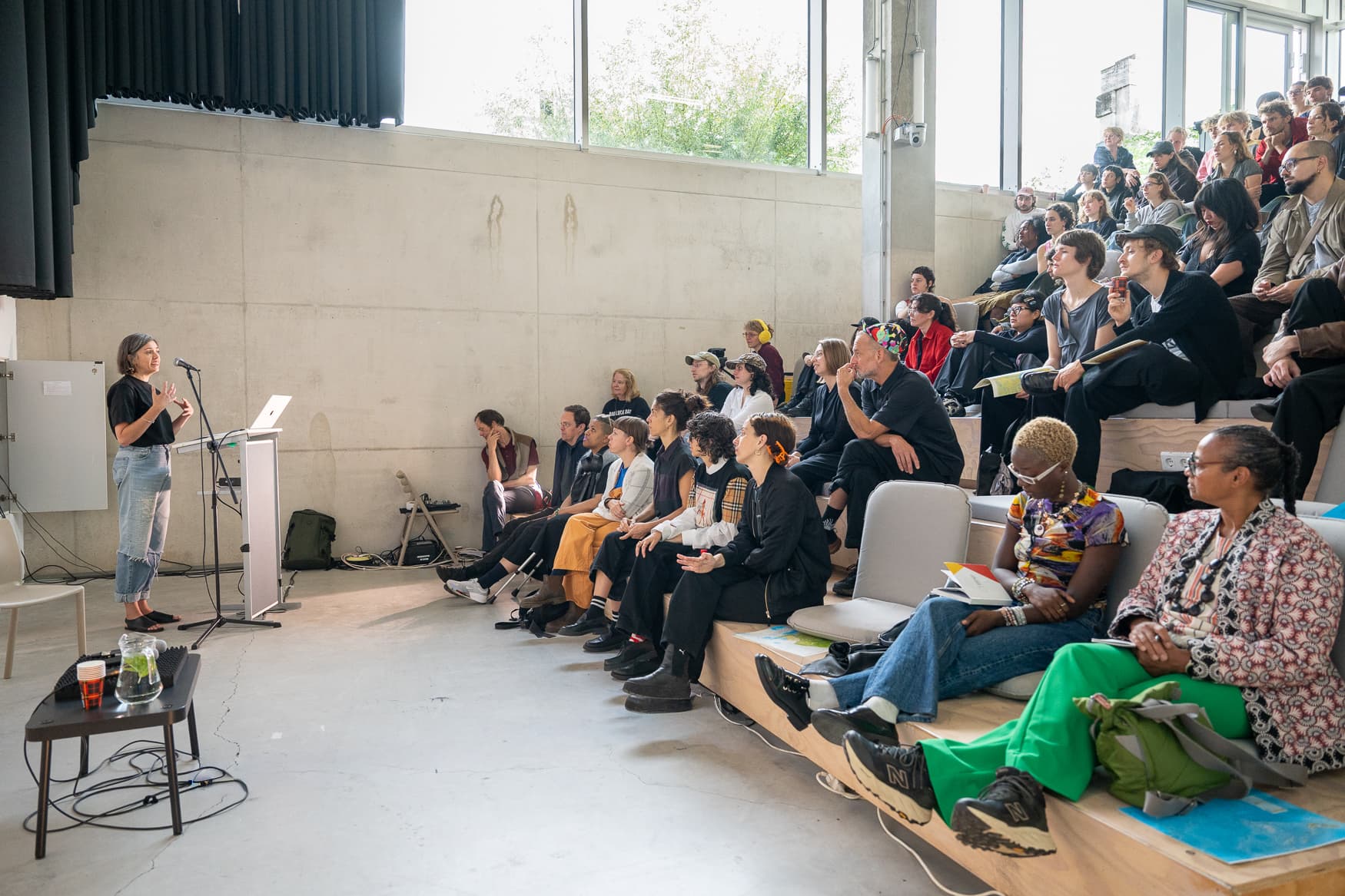 On the left, Ruby Hoette director of the Sandberg Institute is doing a speech looking to the right towards the students sitting in the lecture hall at the academy. The room is brightly lit by the sun shining through large windows.
