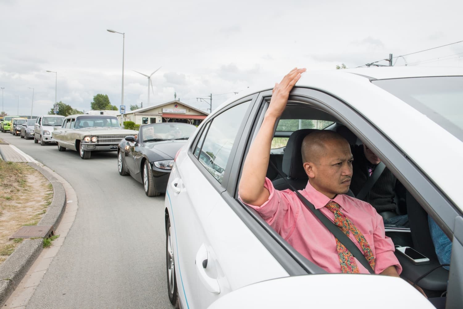 The image shows a person driving a car. They are wearing a pink shirt and the window is open.