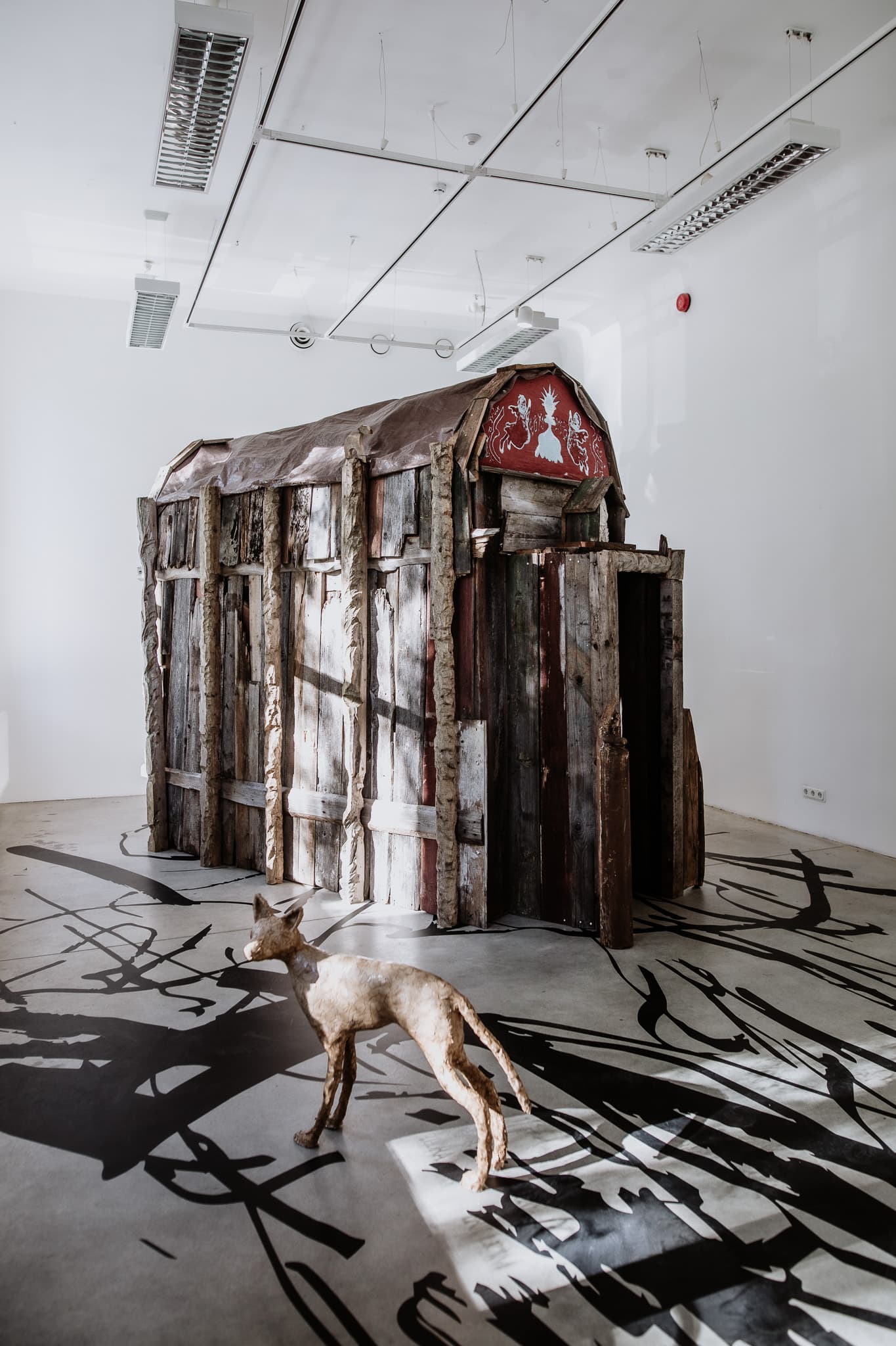 a medium-sized white cube exhibition space. Sunlight coming from large windows illuminate a shabby small chapel built from scavanged wood. The pediment of the buliding features a red sgraffito with angels and other symbols. In the foreground a papier-mache sculpture of a fox-like animal.
