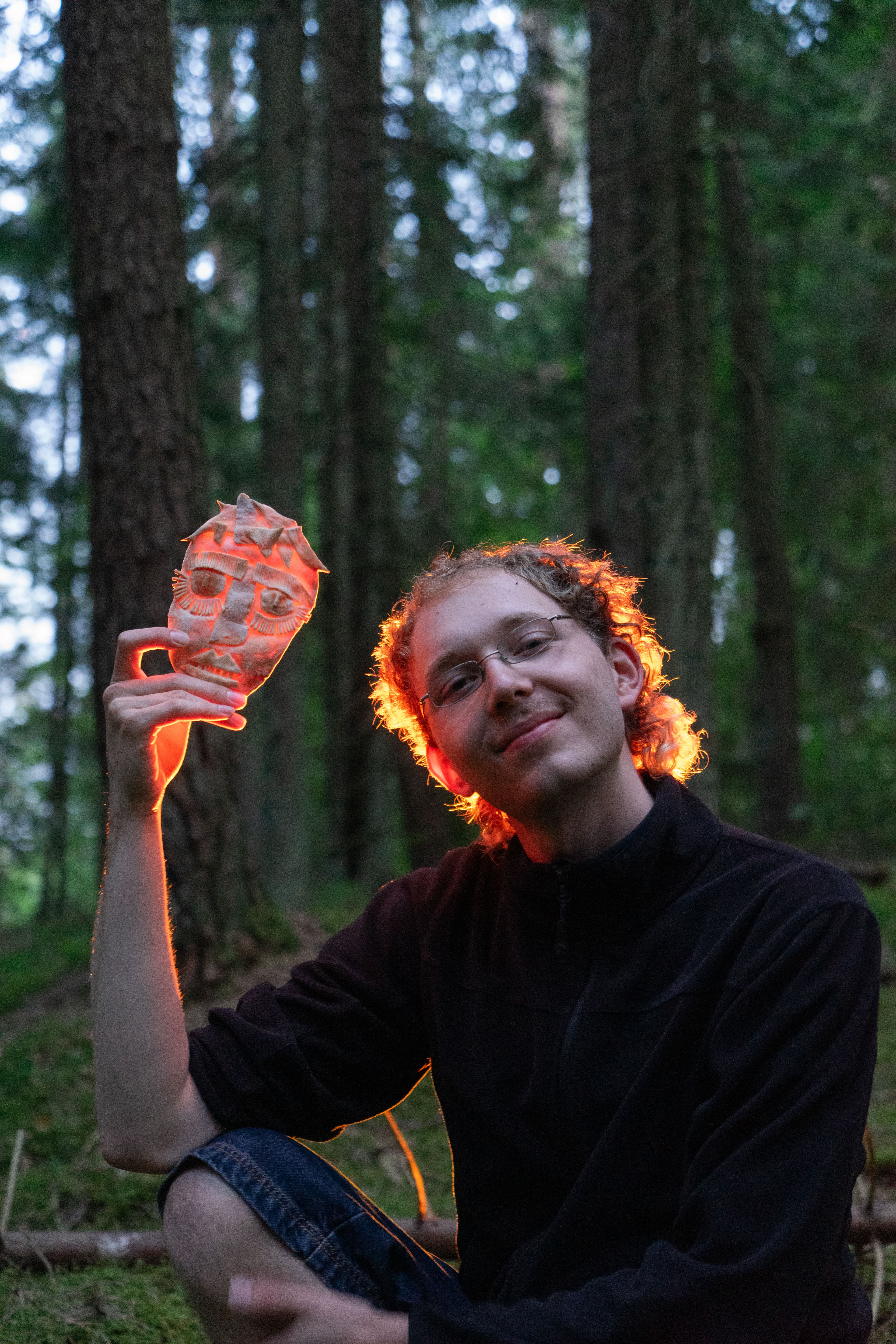 A person sitting on the forest floor with a sculpted mask and light pouring in from the back.