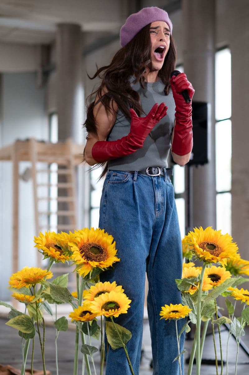 A person is singing into a microphone, surrounded by sunflowers. They have long brow hair, red elbow-length gloves, and a pink beret.